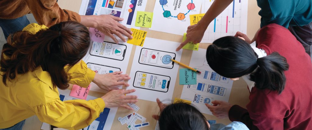 A diverse group of people collaborates around a table, working on a mobile app design project. Printed wireframes, colorful sticky notes, and charts are spread out as team members actively point, write, and discuss ideas. The atmosphere appears creative and focused, emphasizing teamwork and hands-on UX/UI accessibility planning.