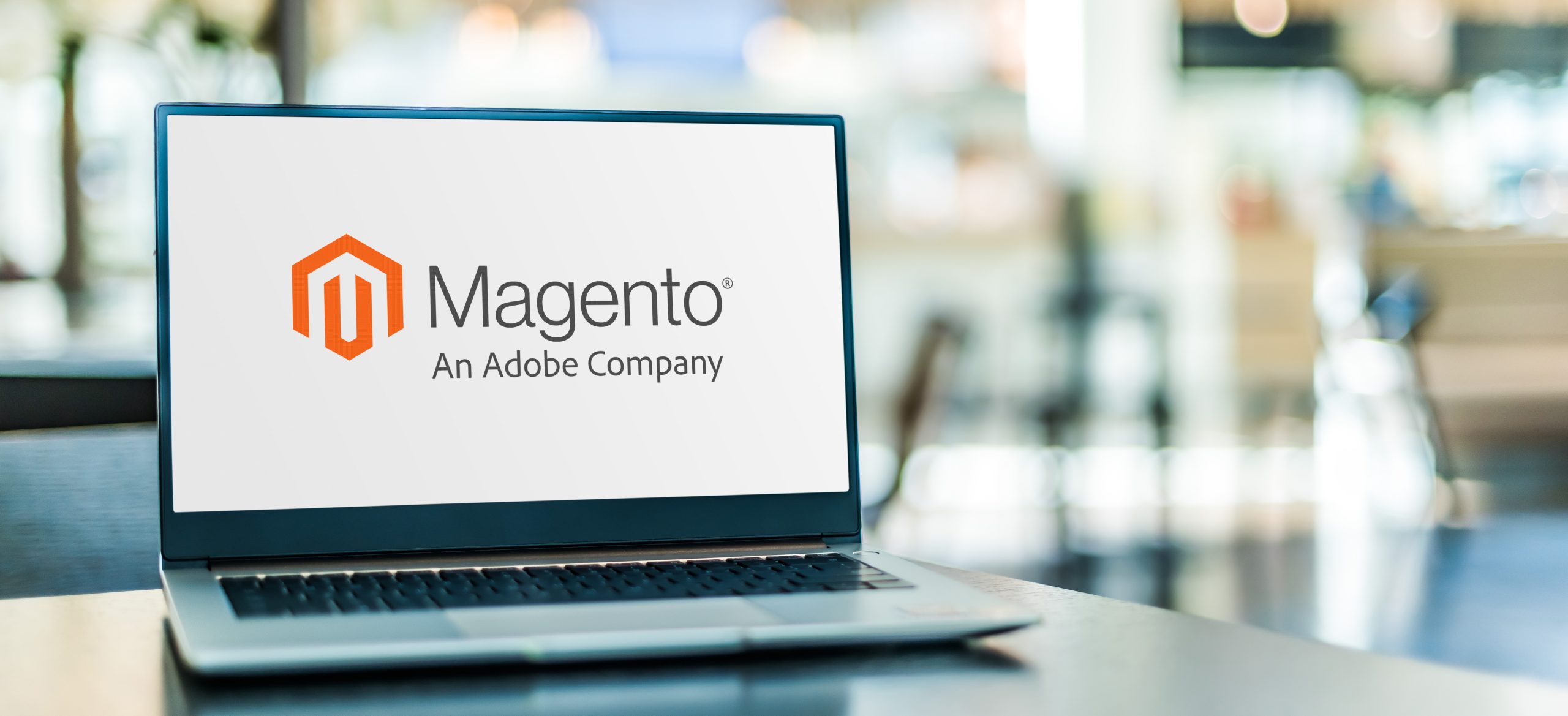 Laptop on a desk displaying the Magento logo with the tagline 'An Adobe Company' on the screen.