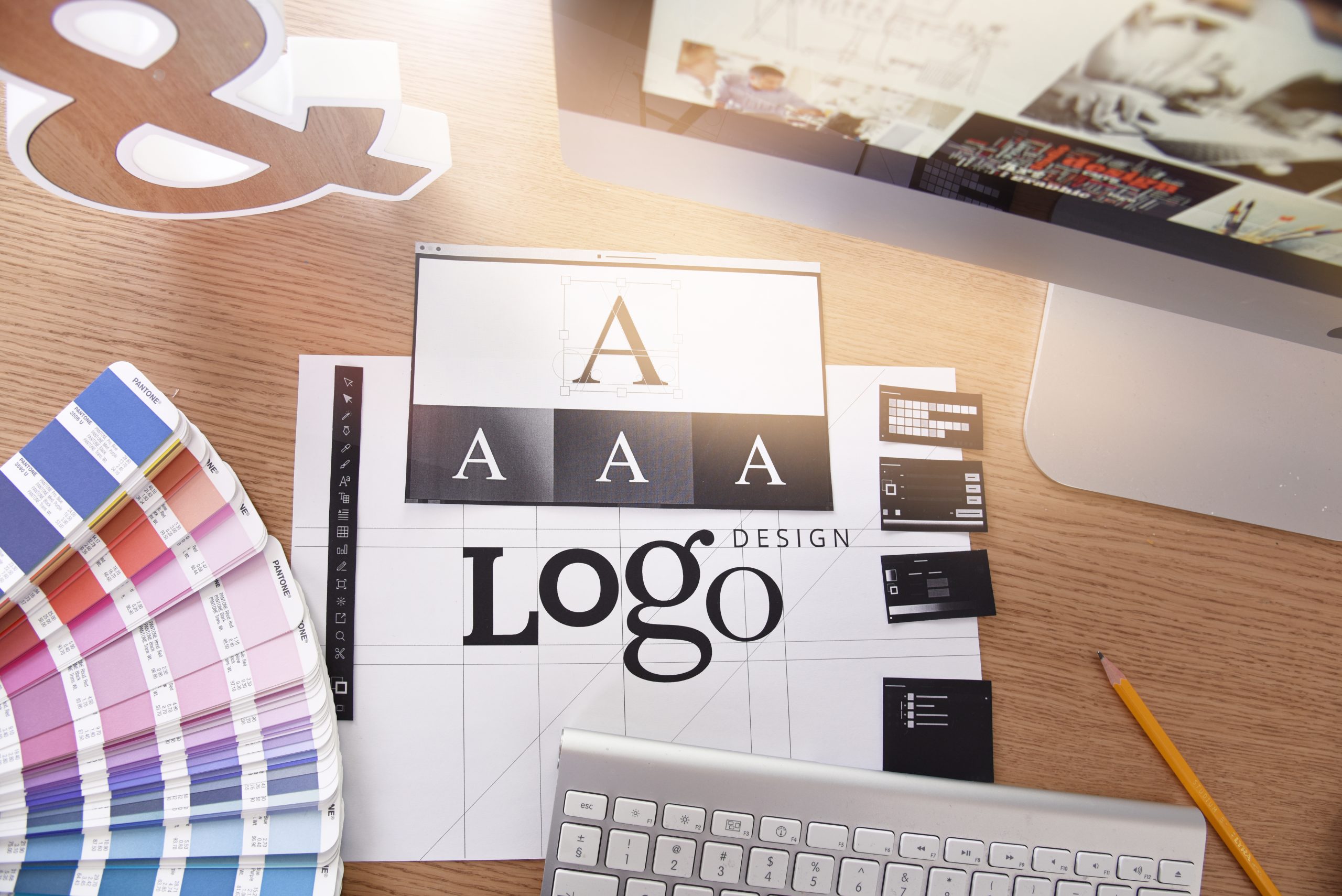 Graphic design workspace flat lay showing branding and logo creation tools, including a Pantone color book, typography sketches, a keyboard, and a computer monitor on a wooden desk.