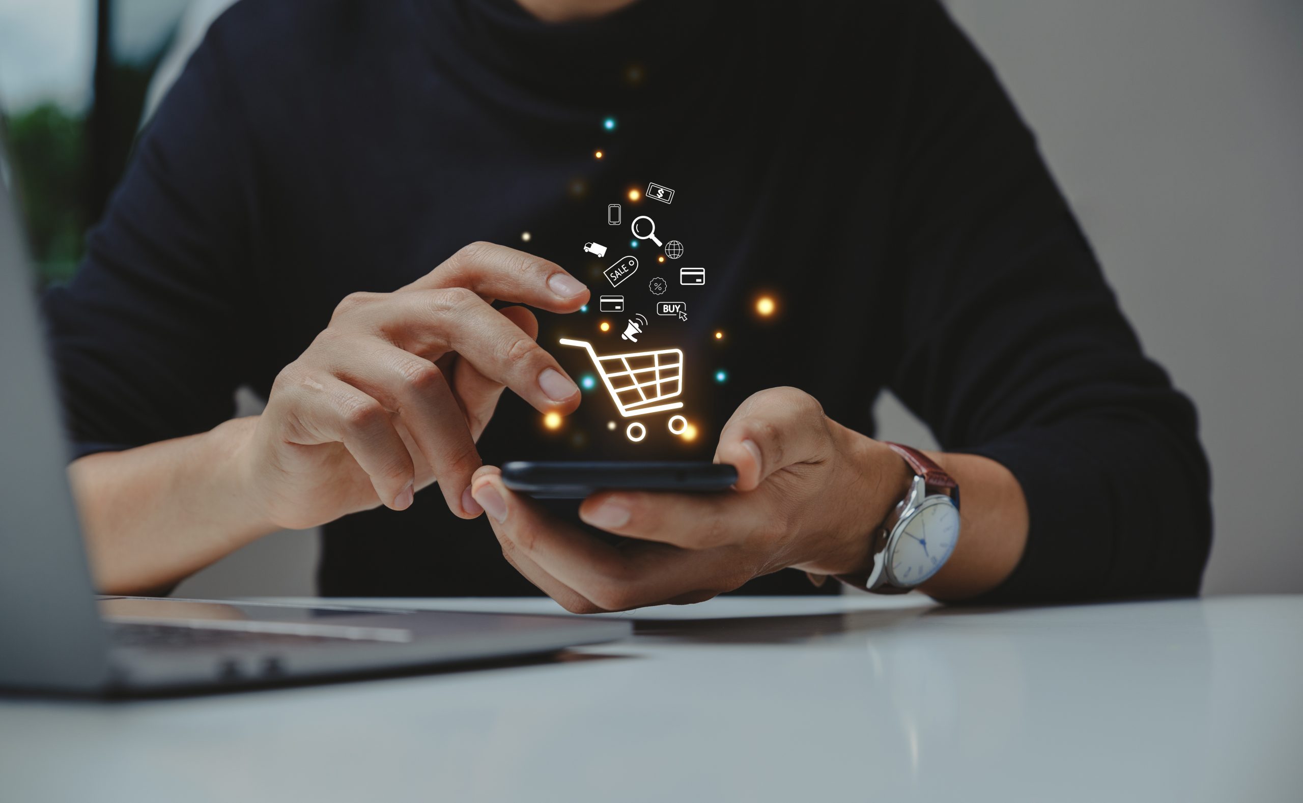 Close-up of a person's hands holding a smartphone, with a glowing digital shopping cart icon and various small e-commerce graphics—like a credit card, delivery truck, and megaphone—floating above the screen. A laptop is partially visible in the foreground on a white desk.