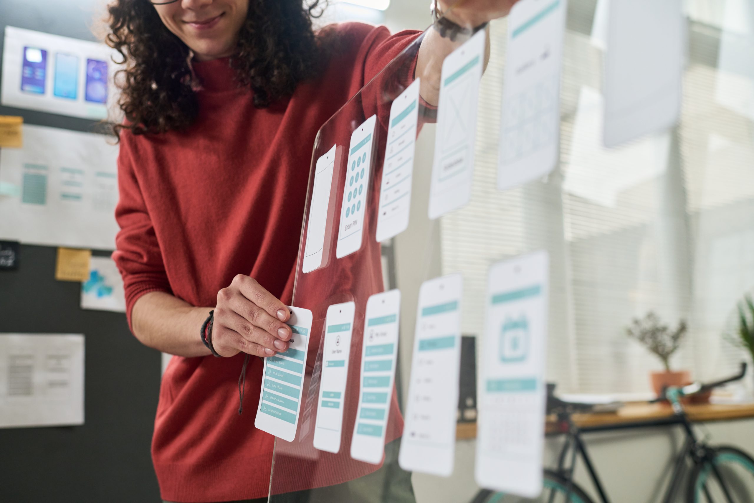 A person with long curly hair wearing a red sweater smiles while arranging paper mobile app interface mockups on a transparent vertical board in a design studio.